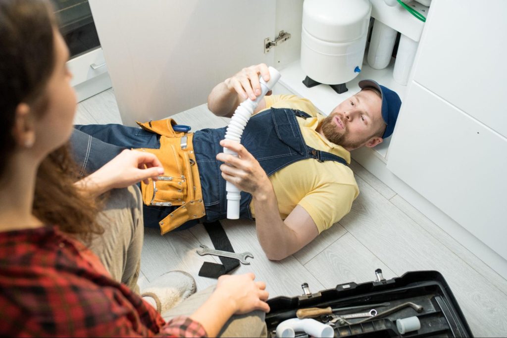 A young plumber replaces the sink trap while the client observes, discussing plumbing prices and service costs.