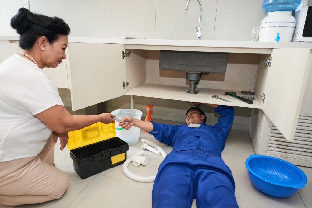 A plumber lying on the ground is fixing the pipes while an older woman is assisting him.