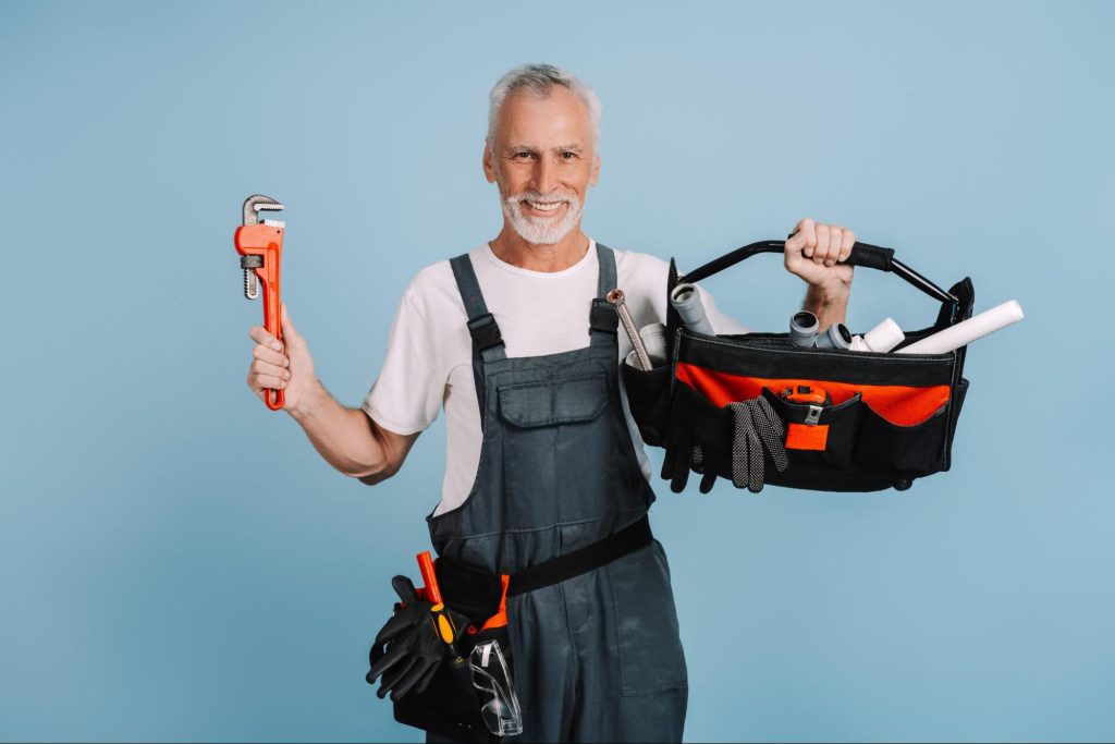A smiling, bearded local plumber wearing a white shirt under a denim jumper holds a pipe wrench and tool bag against a blue background.