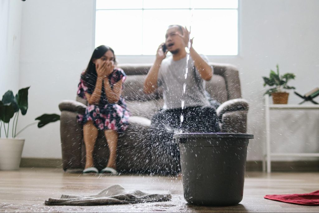 A worried man on the phone calls for help while his frustrated wife looks at a severe roof leak, signaling a potential plumbing emergency causing water damage in their home.
