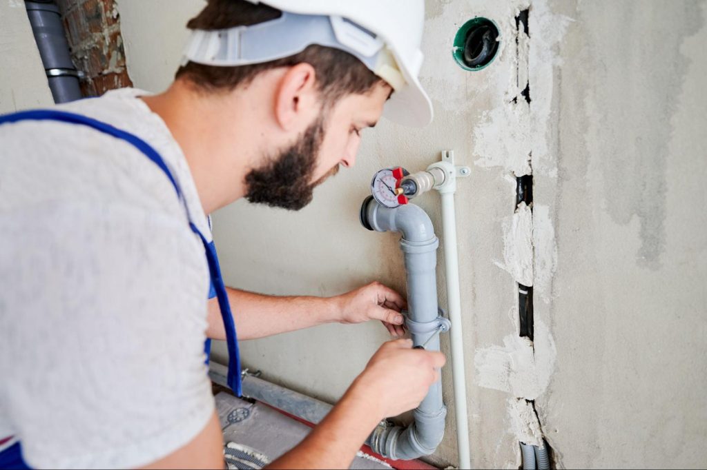A male plumber in a hardhat screwed a clamp on a pipe with a screwdriver.