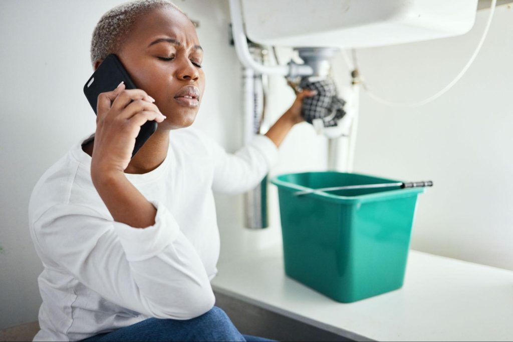 A woman holding a leaking pipe under the sink calls a plumbing company for help.