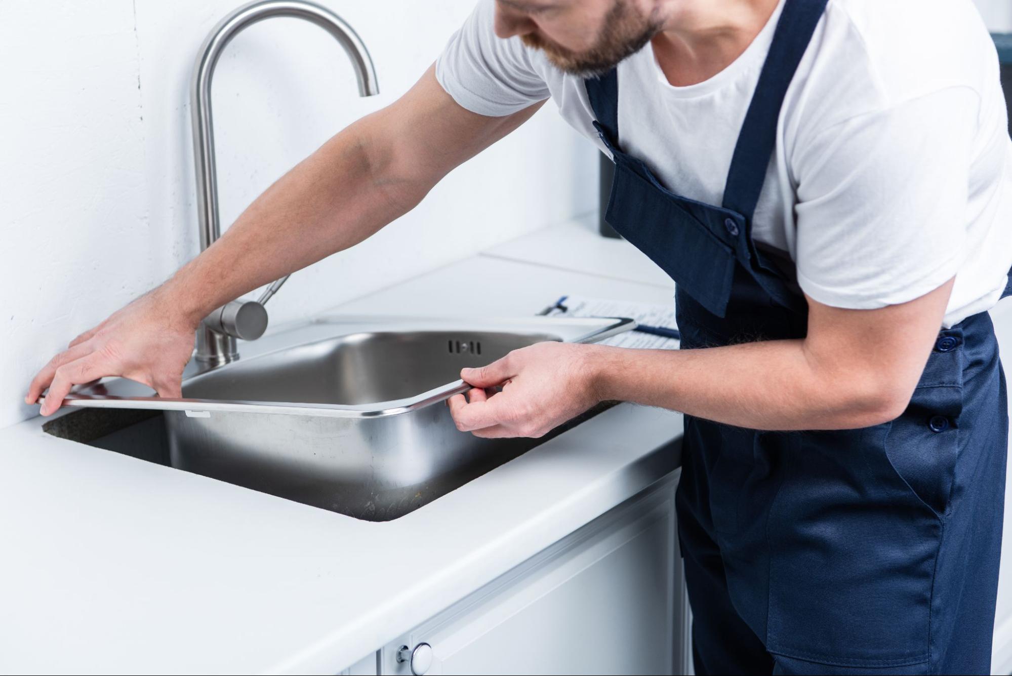 Partial view of a bearded repairman in overalls doing sink repair in the kitchen.