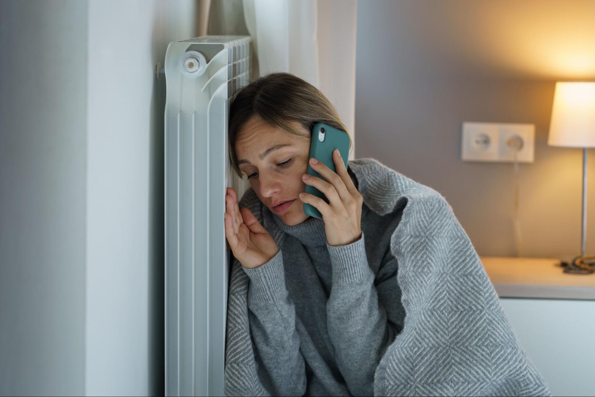 A young woman calls an emergency heating services while leaning on the radiator.