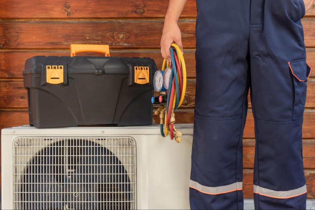 An HVAC technician holding a manifold gauge stands beside an outdoor condenser unit with an equipment bag resting on top.